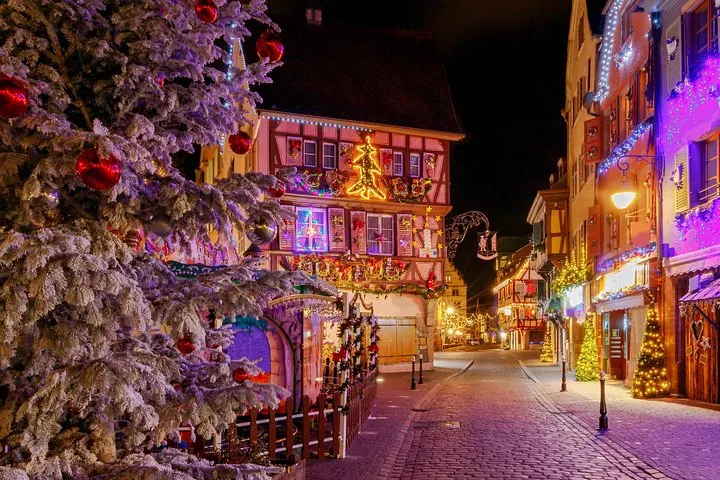 A snow-dusted Christmas tree in front of a half-timbered street lit in pink and violet at night in Colmar