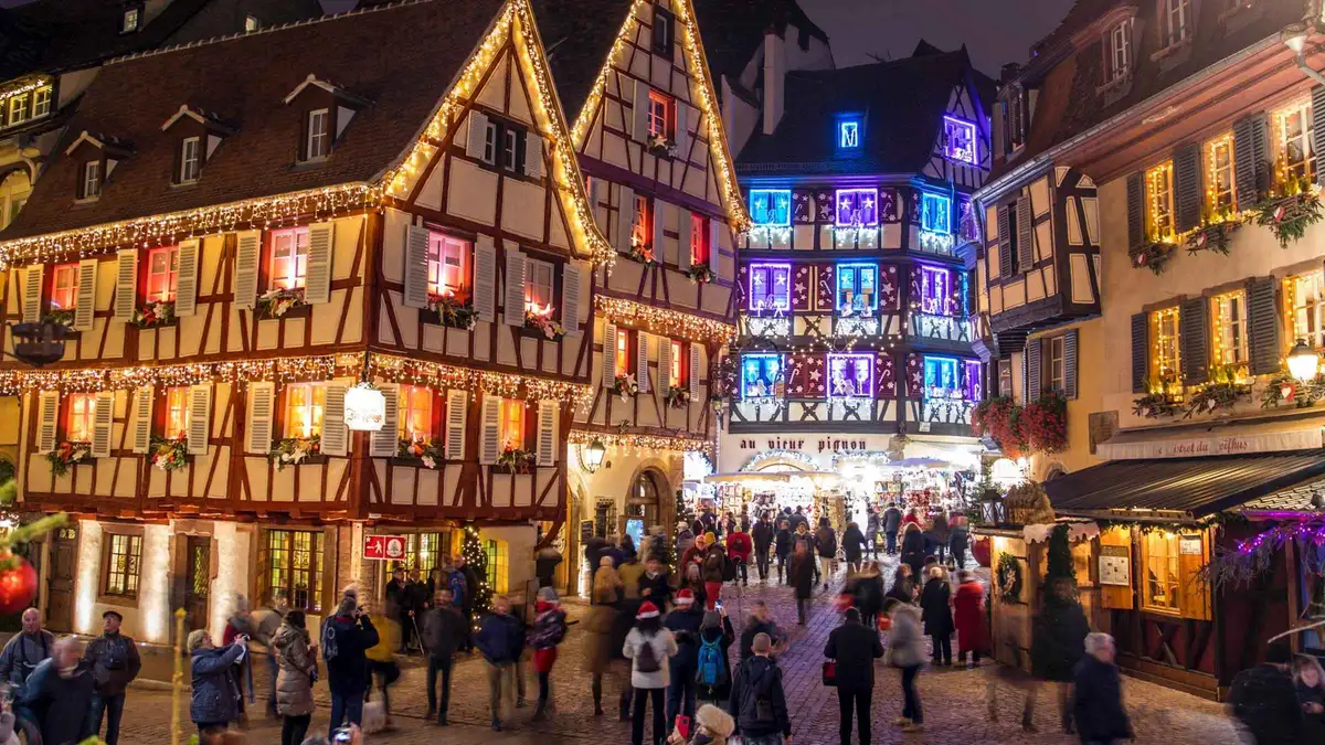 Visitors walking a lit street of half-timbered houses dressed for Christmas in Colmar at dusk