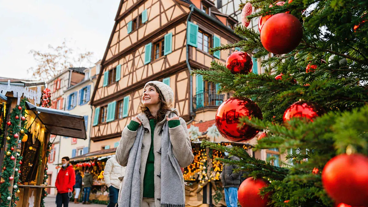 A woman in a white coat looking up at red baubles on a Christmas tree beside a market stall in Colmar