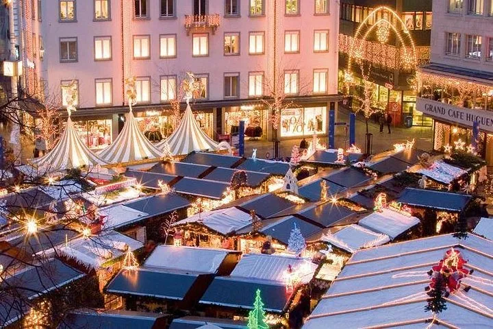 White-roofed Christmas market stalls packed into a city square at dusk, with lit hotel facades behind