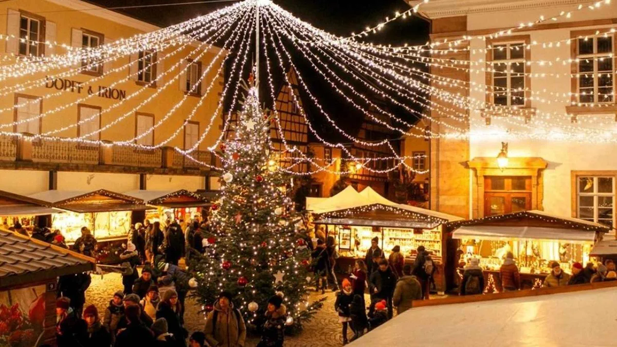 A lit Christmas tree and market huts under strings of lights crossing a village square at night, crowds between the stalls